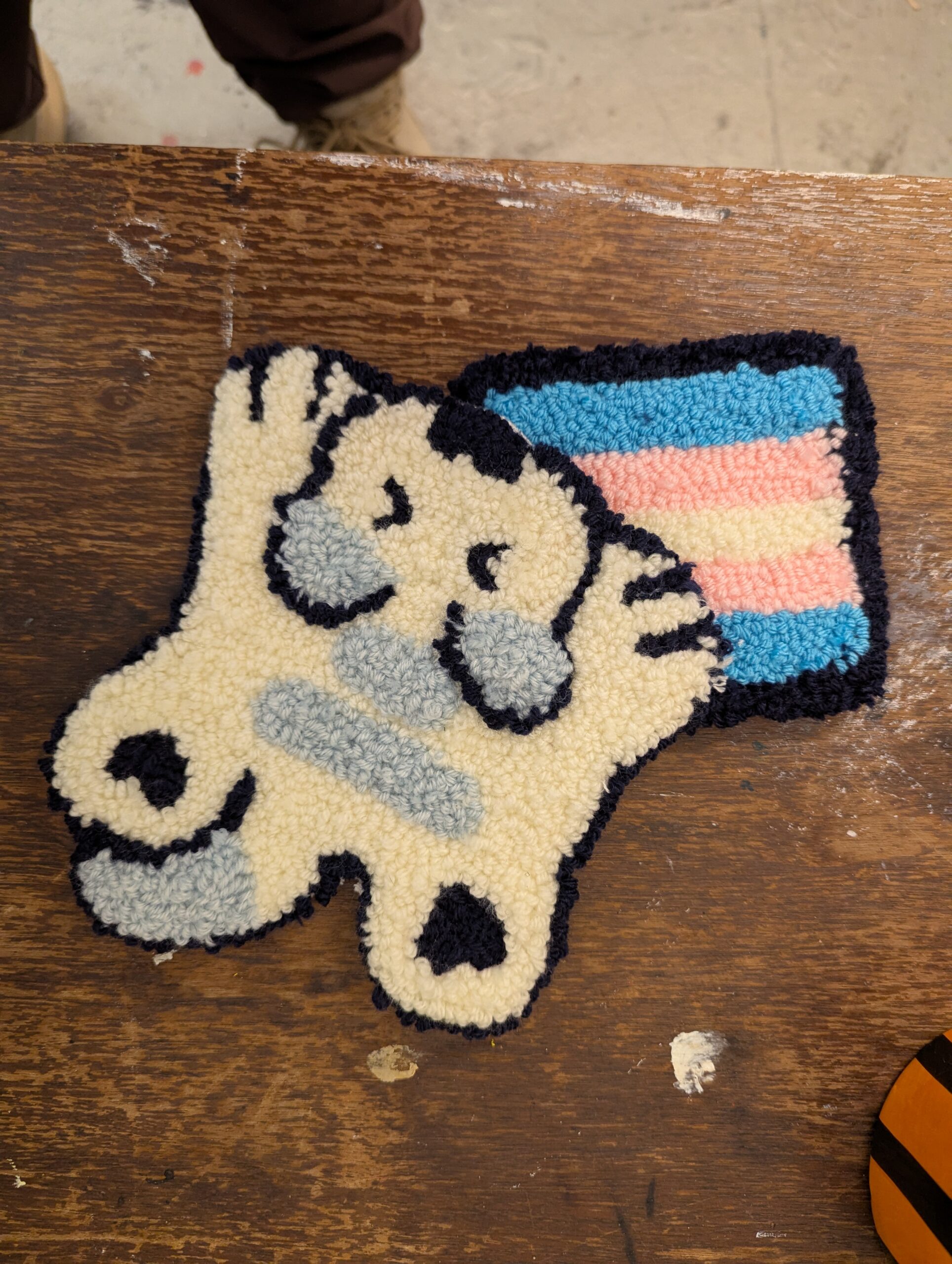 A photo of two rugs. The bottom is a trans flag with a dark border. The top is a white and grey cat with its arms and legs sprawled out
