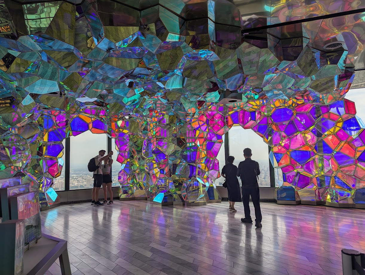 A metal arch of assymetric geometric shapes stretching from floor to ceiling. The faces of the structure have a holographic plexiglass plate that reflects light into many colors. Through the arch you can see the windows of Hudson Yards, with a view of NYC from a hundren stories up and the water.